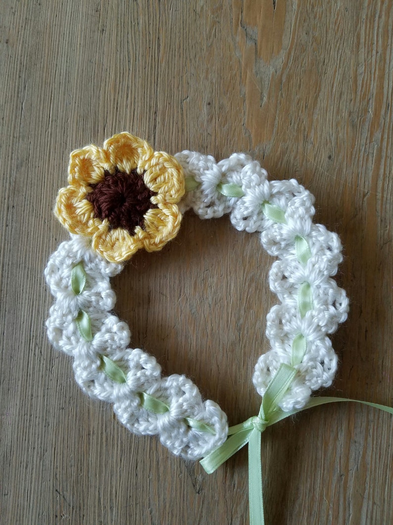 May include: A white crocheted headband with a green ribbon tied at the back. A yellow crocheted sunflower with a brown center is attached to the headband.