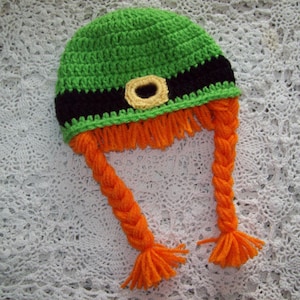 Puede incluir: Sombrero de ganchillo verde con una banda negra y una hebilla dorada. El sombrero tiene dos trenzas largas de color naranja.
