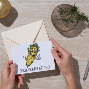May include: A white card with a cartoon corn on the cob with a smiling face and thumbs up. The text on the card reads "Corn-gratulations!"