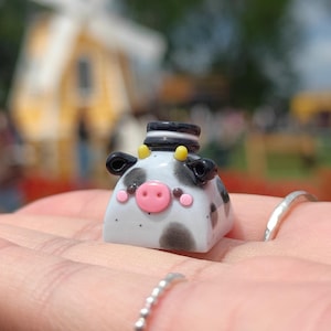 Puede incluir: Una pequeña figura de vaca cuadrada con manchas blancas y negras, una nariz rosa y un sombrero de copa blanco y negro. La vaca tiene pequeñas orejas negras y cuernos amarillos. La figura se sostiene en una mano, con un molino de viento al fondo.