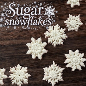 Könnte beinhalten: Nahaufnahme von weißen Zuckerschneeflocken auf dunklem Holzgrund. Das Bild zeigt mehrere detaillierte, essbare Schneeflocken mit aufwendigen Designs. Der Text "Sugar snowflakes" ist in weißer Schrift dargestellt, was auf ein Winter- oder Feiertagsmotiv hindeutet.