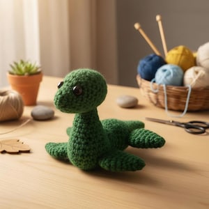 May include: A handmade green crochet sea monster with large black eyes. The creature is lying on a wooden surface. Knitting supplies, including yarn and needles, are in the background, suggesting a craft project.