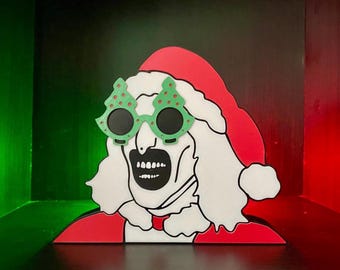 May include: A festive Christmas decoration featuring a stylized Santa Claus figure with a unique twist. The figure wears a red Santa hat and suit, with white hair and a grimacing expression. The figure is wearing green Christmas tree-shaped glasses.