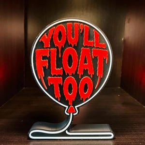 May include: A black and red balloon-shaped sign with the text "YOU'LL FLOAT TOO" in dripping red letters. The sign is mounted on a black base with a white outline, and is a decorative item.
