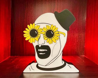 May include: A white, black, and yellow decorative figure of a clown-like face with sunflower-shaped glasses and a small black hat. The figure has a black collar and a wide open mouth.