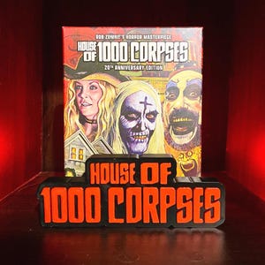 May include: A black and orange "House of 1000 Corpses" sign sits in front of a DVD case. The DVD case features a movie poster with three characters. The text "House of 1000 Corpses" and "20th Anniversary Edition" are visible.