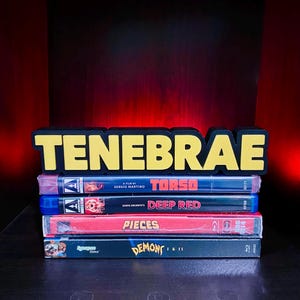 May include: A stack of horror film Blu-ray and DVD cases, including "Torso," "Deep Red," "Pieces," and "Demons 1 & 2." The word "TENEBRAE" is displayed in yellow block letters above the cases.