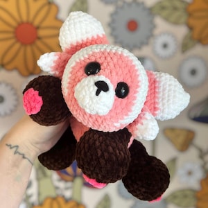 May include: A handmade crochet red panda plushie. The plushie features a pink and white head with black eyes and a white muzzle. The ears and tail tips are white, and the paws are brown with pink accents. The plushie is held in a hand.