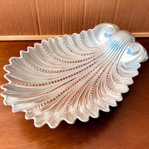 Puede incluir: Un plato de metal plateado con forma de concha marina grande. El plato presenta un borde festoneado y una serie de crestas elevadas y radiantes que se extienden desde el área de la bisagra hacia el borde exterior, creando un aspecto texturizado.
