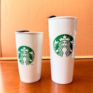 May include: Two white ceramic travel mugs of different heights, each featuring the green and white Starbucks siren logo on the front and a black lid. The mugs have a smooth, cylindrical body and a tapered base, displaying the iconic brand emblem.