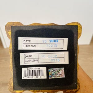 Pu&ograve; includere: Primo piano della base di una lampada decorativa con una cornice dorata e ornata. La base ha un fondo in feltro nero con due etichette bianche che mostrano la data "10 2002" e il numero articolo "10215". &Egrave; visibile anche un adesivo con informazioni di sicurezza.