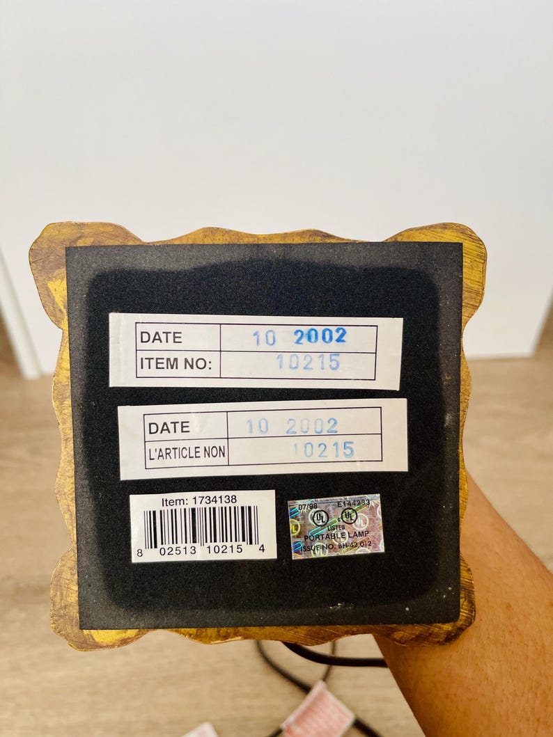 Pu&ograve; includere: Primo piano della base di una lampada decorativa con una cornice dorata e ornata. La base ha un fondo in feltro nero con due etichette bianche che mostrano la data "10 2002" e il numero articolo "10215". &Egrave; visibile anche un adesivo con informazioni di sicurezza.