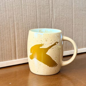 May include: A light yellow ceramic mug featuring a gold silhouette of a flying goose on the exterior. The mug has a speckled finish and the text Tim Hortons is printed in gold on the interior rim. It includes a curved handle on the right side.