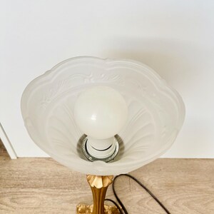Pu&ograve; includere: Una lampada da tavolo vintage con base color oro e paralume in vetro smerigliato. Il paralume ha un bordo smerlato e un motivo floreale. La lampada ha una lampadina standard e un cavo di alimentazione marrone. La lampada &egrave; su una superficie di legno.