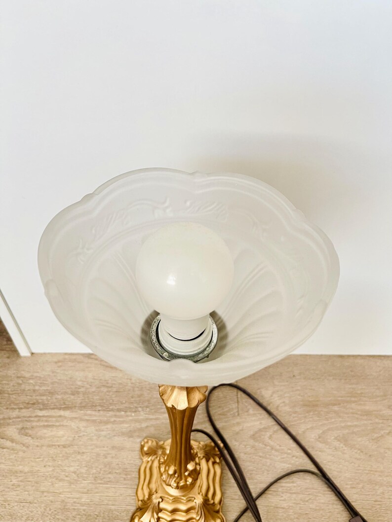 Pu&ograve; includere: Una lampada da tavolo vintage con base color oro e paralume in vetro smerigliato. Il paralume ha un bordo smerlato e un motivo floreale. La lampada ha una lampadina standard e un cavo di alimentazione marrone. La lampada &egrave; su una superficie di legno.