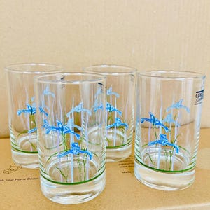 Può includere: Quattro bicchieri trasparenti con un motivo floreale. Ogni bicchiere presenta un motivo di iris blu con steli e foglie verdi. I bicchieri sono cilindrici e poggiano su una superficie di cartone.