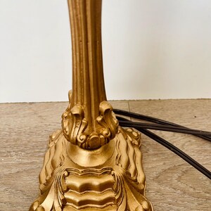 Pu&ograve; includere: Una base per lampada color oro con dettagli classici ornati. La base ha un design a colonna scanalata e una base quadrata decorativa con accenti a volute. Cavo elettrico marrone visibile.