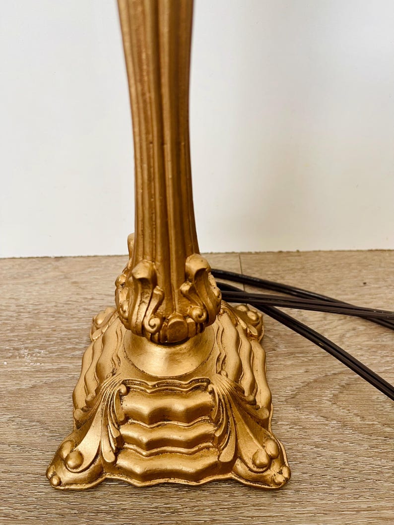 Pu&ograve; includere: Una base per lampada color oro con dettagli classici ornati. La base ha un design a colonna scanalata e una base quadrata decorativa con accenti a volute. Cavo elettrico marrone visibile.