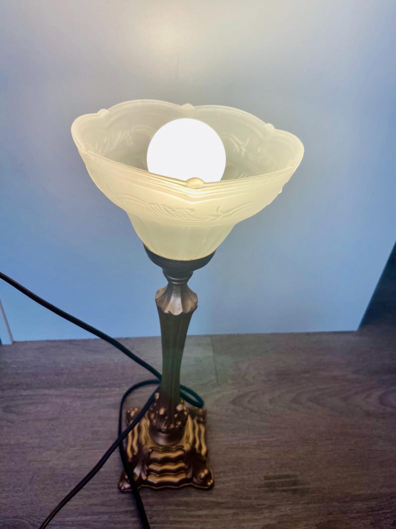 Pu&ograve; includere: Una lampada da tavolo in stile antico con un paralume in vetro smerigliato e una base decorativa. Il paralume presenta un motivo floreale e circonda una lampadina luminosa. La base &egrave; ornata con una finitura bronzo. La lampada &egrave; collegata con un cavo nero.
