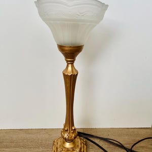 Pu&ograve; includere: Una lampada da tavolo in stile vintage con base e stelo color oro. La lampada presenta un paralume in vetro smerigliato con un motivo floreale decorativo. La base ha un design ornato e dettagliato. La lampada &egrave; collegata con un cavo nero.