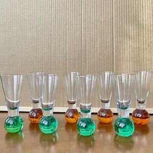 以下が含まれることがあります： 丸いベースが付いた透明なガラスのショットグラスのセット。ベースは緑色とオレンジ色を交互に配置。ショットグラスは木製の表面に一列に並んでいます。