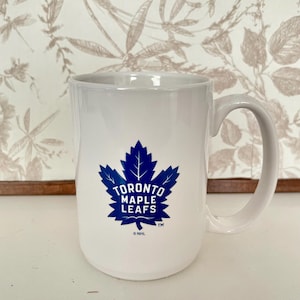 Puede incluir: Taza de cerámica blanca con el logotipo de los Toronto Maple Leafs en azul. El logotipo incluye una hoja de arce estilizada y el nombre del equipo. La taza tiene un asa curva y es adecuada para bebidas frías o calientes.