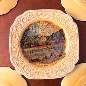May include: A collection of decorative plates with a cream-colored border and a central image of an English river scene. The plates have a scalloped edge and are arranged on a dark wooden surface. The text "English River Scenes" is visible.