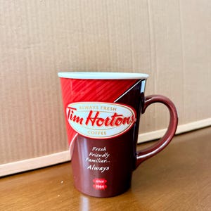 May include: A ceramic mug featuring a red and dark brown colour scheme with diagonal stripes. The front displays the Tim Hortons logo in red text inside an oval, with the words Always Fresh, Fresh Friendly Familiar... Always, and since 1964.
