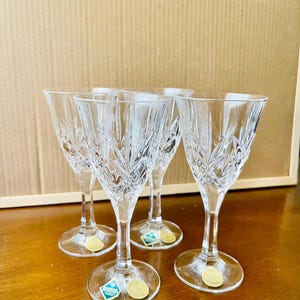 Puede incluir: Cuatro copas de vino de cristal transparente con intrincados diseños grabados. Cada copa tiene un tallo delgado y una base redonda. Pequeñas pegatinas azules y doradas están adheridas a la base de cada copa. Las copas están sobre una superficie de madera.