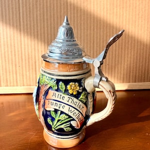 May include: A decorative beer stein with a silver metal lid and handle. The stein features a blue and brown design with the words "Alte Thaler Junge Weiber" on a banner. The handle is cream-coloured.