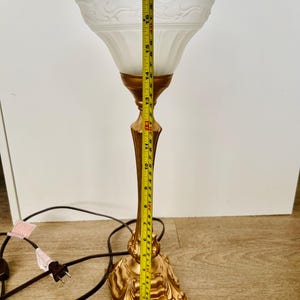 Pu&ograve; includere: Una lampada da tavolo vintage con base e stelo color oro e paralume in vetro smerigliato bianco. La lampada &egrave; alta circa 46 cm. Il paralume presenta un motivo floreale decorativo. Un cavo di alimentazione nero si estende dalla base.