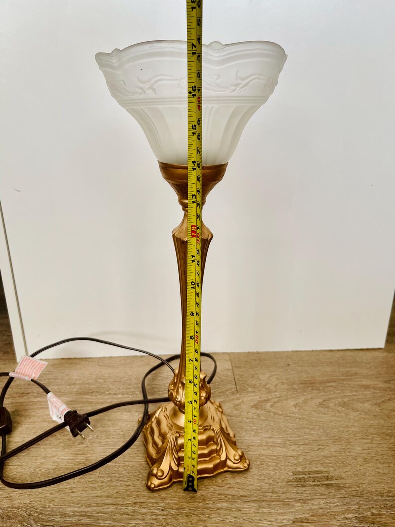 Pu&ograve; includere: Una lampada da tavolo vintage con base e stelo color oro e paralume in vetro smerigliato bianco. La lampada &egrave; alta circa 46 cm. Il paralume presenta un motivo floreale decorativo. Un cavo di alimentazione nero si estende dalla base.