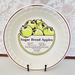 May include: A white ceramic pie dish with a scalloped edge and a red border. The dish displays a graphic of green apples and the text "Sugar Bread Apples" with a recipe.