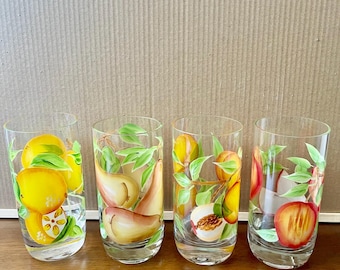 Könnte beinhalten: Vier klare Trinkgläser mit handgemalten Fruchtdesigns. Jedes Glas zeigt eine andere Frucht: Zitronen, Birnen, Pfirsiche und Pflaumen, mit grünen Blättern. Die Gläser sind auf einer Holzoberfläche angeordnet.