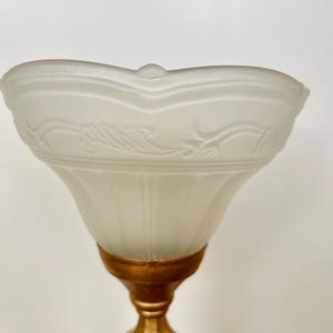Pu&ograve; includere: Primo piano di una lampada vintage. La lampada presenta un paralume in vetro smerigliato con un design floreale ornato e una base dorata. Il paralume ha un bordo smerlato e un piccolo dettaglio arrotondato sulla parte superiore.