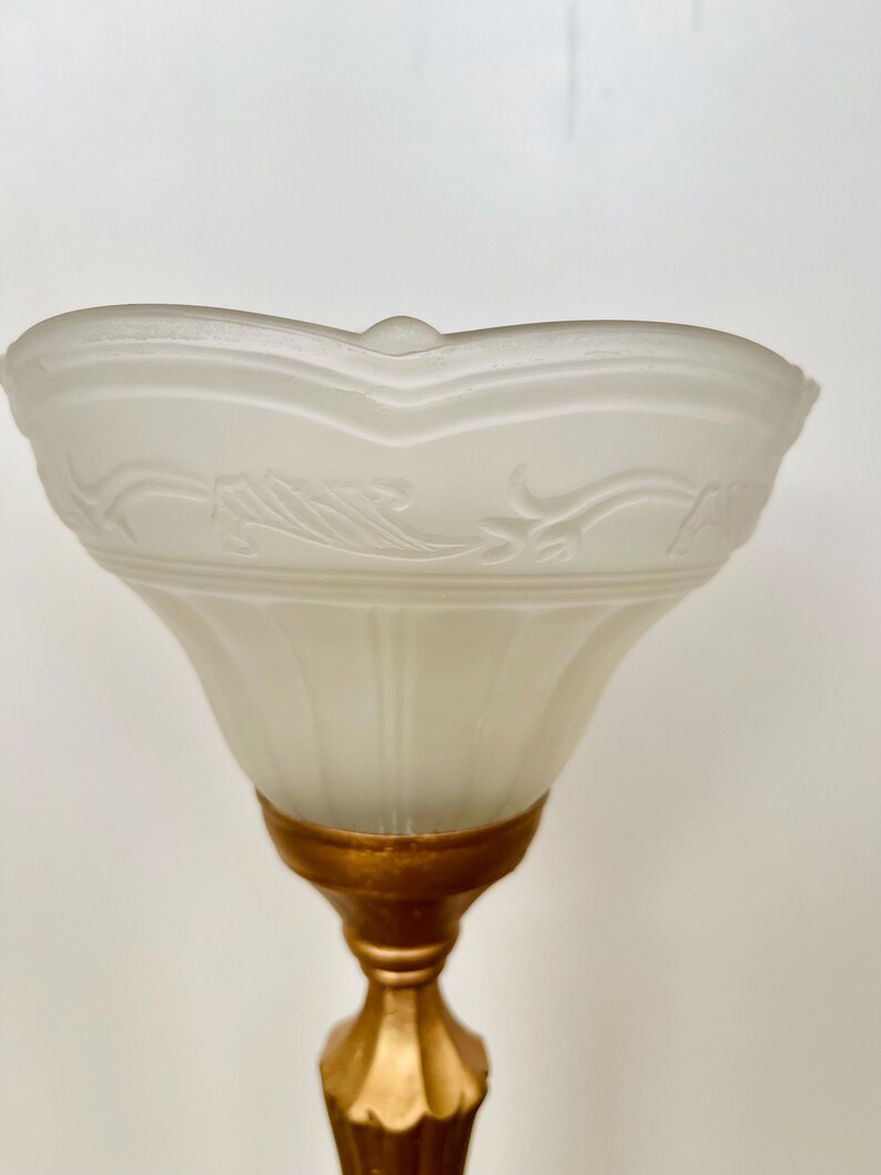 Pu&ograve; includere: Primo piano di una lampada vintage. La lampada presenta un paralume in vetro smerigliato con un design floreale ornato e una base dorata. Il paralume ha un bordo smerlato e un piccolo dettaglio arrotondato sulla parte superiore.