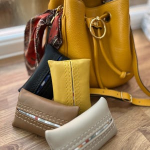 May include: Four different colored leather tissue holders with decorative stitching. The holders are arranged in a row on a wooden surface. A yellow leather bucket bag is in the background.