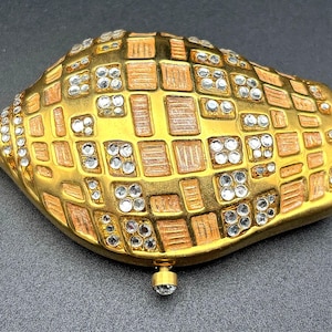 May include: A gold-toned, shell-shaped decorative object with a mosaic-like pattern. The squares are a mix of gold and light brown, with clear, sparkling crystals embedded throughout. A small, crystal-tipped clasp is visible at the bottom.