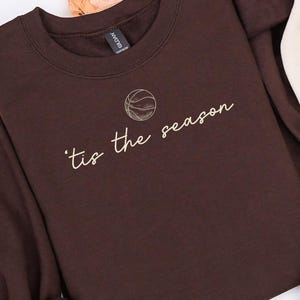 May include: Two sweatshirts, one brown and one cream, with the text "'tis the season" and a basketball graphic. The sweatshirts are long-sleeved and have a crew neck design. Autumn leaves are in the background.