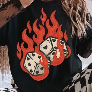 May include: Black graphic t-shirt featuring two dice engulfed in orange flames. The dice have heart-shaped pips. The person is wearing a white and red hat with the word "TIMELESS" and black and white checkered pants.