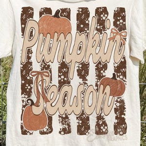 May include: A cream-colored short-sleeve t-shirt featuring a graphic print of brown vertical stripes with a white floral pattern. The text "Pumpkin Season" is written in large beige script, accompanied by three orange pumpkins and brown bows.
