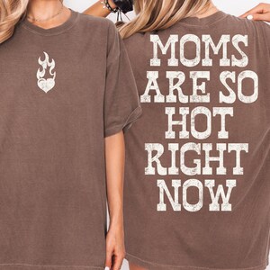 May include: Brown t-shirt with the text "MOMS ARE SO HOT RIGHT NOW" on the back. The front of the shirt features a flame heart graphic. The shirt has short sleeves and a relaxed fit.