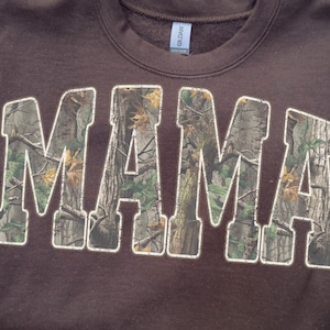 May include: A brown sweatshirt with the word "MAMA" in large, block letters. The letters are filled with a camouflage pattern in shades of green, brown, and tan. The sweatshirt has a crew neck and long sleeves.