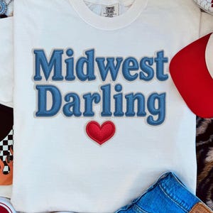 Puede incluir: Una camiseta color crema con el texto bordado "Midwest Darling" en azul, un corazón rojo y una gorra de béisbol roja y blanca. La imagen también incluye vaqueros azules y zapatillas rojas.