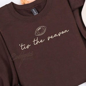 May include: A dark brown sweatshirt and a cream-colored sweatshirt, both featuring a graphic of a football and the text 'tis the season in a cursive font. The garments have crew necklines and long sleeves, presented as a pair of matching tops.