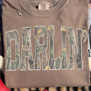 Puede incluir: Una sudadera marrón Comfort Colors con la palabra "DARLIN'" en blanco, con un diseño de camuflaje. Un collar con una cruz dorada descansa sobre la camisa. La camisa está doblada sobre una superficie con piel de vaca e impresión animal.