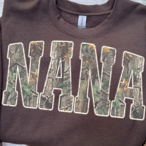 May include: A brown sweatshirt with the word "NANA" in large, block letters. The letters are filled with a camouflage pattern and outlined in white. The sweatshirt is a solid color and has a crew neck.