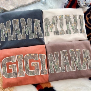 May include: Four sweatshirts in various colors: charcoal, beige, rust, and brown. Each sweatshirt features a different word in a camouflage print: "MAMA", "MINI", "GIGI", and "NANA". The sweatshirts are folded and stacked.