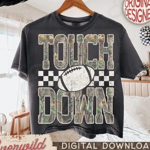 May include: A charcoal gray t-shirt featuring a graphic design with the text TOUCH DOWN in a camouflage pattern. A white football illustration is centered between the words, and a black and white checkered border accents the design.
