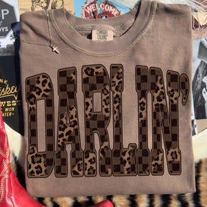 Op de afbeelding: Een bruine Comfort Colors sweatshirt met het woord "DARLIN'" in grote, vetgedrukte letters. De letters hebben een dambord- en luipaardprint ontwerp. Een gouden ketting met een kruisvormige hanger is zichtbaar.